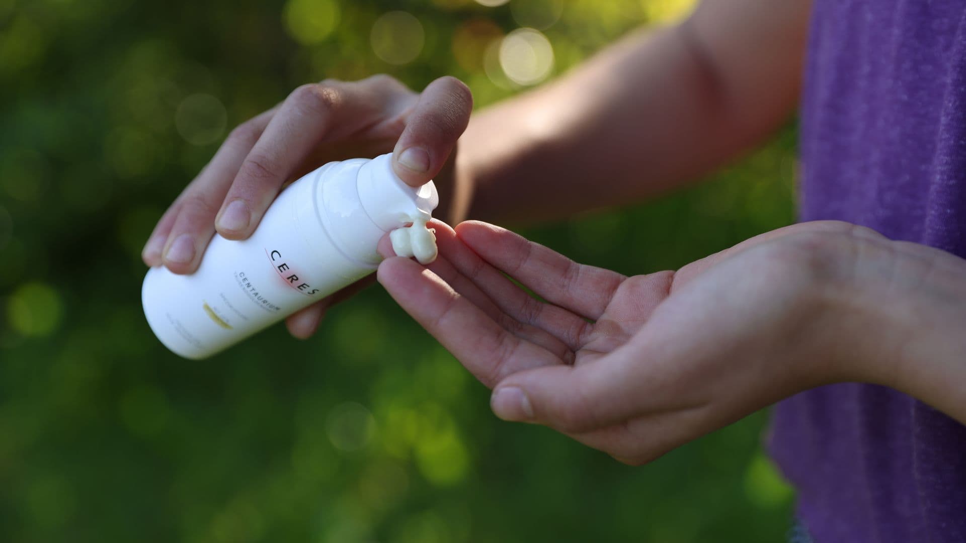 Person drückt eine weiße Lotion aus einer Flasche mit der Aufschrift 'CERES' auf die offene Hand vor einem unscharfen grünen Hintergrund.