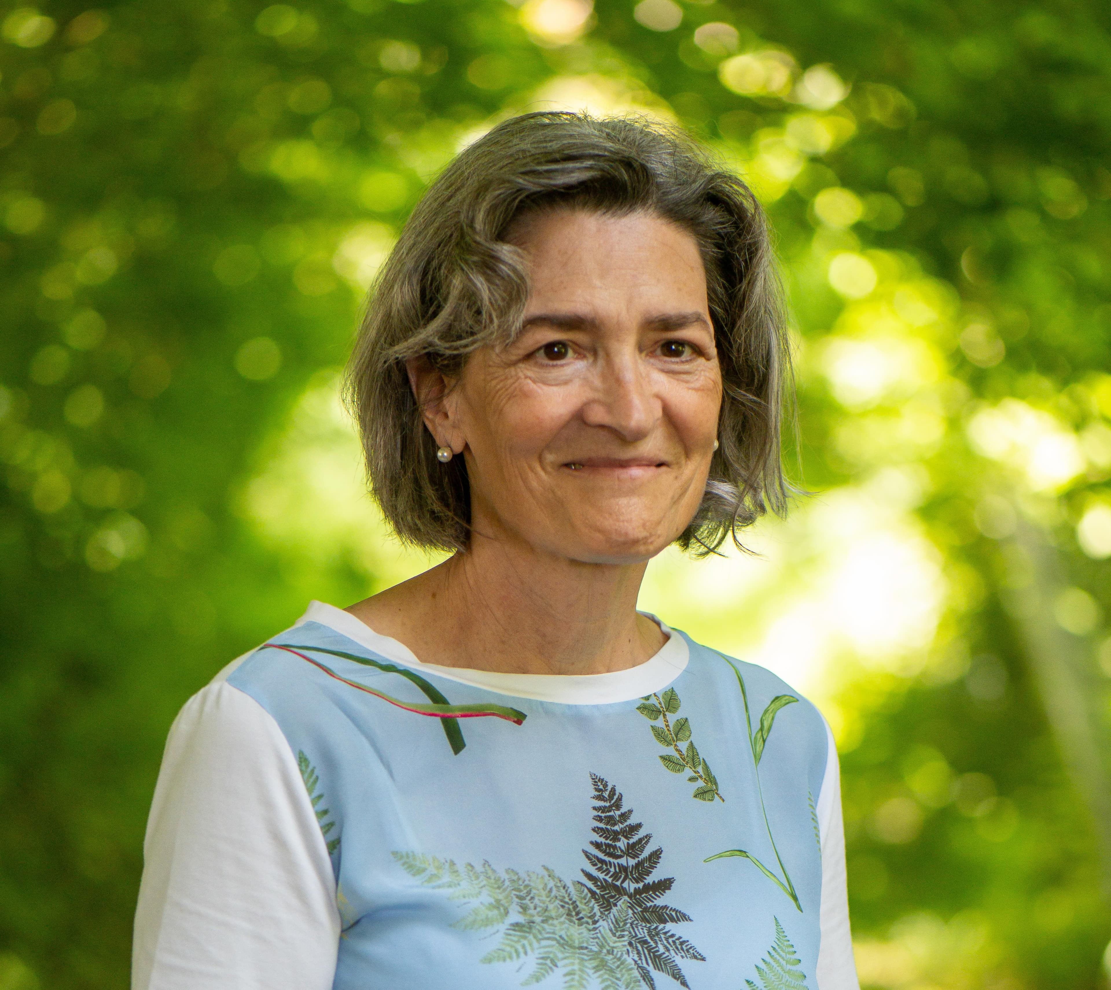 Frau mit kurzem grauem Haar trägt ein hellblaues T-Shirt mit weißen Ärmeln und bunten Pflanzenmotiven, vor unscharf grünem Hintergrund im Freien.